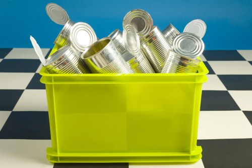 A bright yellow plastic container filled with numerous empty, silver tin cans, some with open tops and others with sealed lids, is positioned on a black and white checkered floor. The background features a plain blue wall, suggesting an indoor setting possibly within a waste collection or recycling area. The cans are stacked haphazardly, with some leaning and others upright, indicating they are prepared for removal or recycling. The container is rectangular with smooth surfaces and rounded edges, and the overall scene appears clean and organized, reflecting a typical rubbish collection process overseen by Bow House Clearance in the Bow area when handling metal waste or recyclable materials.