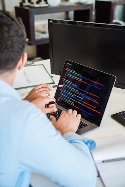 A person wearing a light blue long-sleeved shirt is seated at a white desk, working on a laptop that displays colorful lines of computer programming code on its screen. The person's left hand is holdi