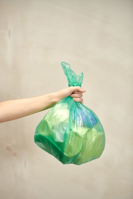 A person's hand and forearm extend into the frame from the left side, grasping a green plastic rubbish bag by its twisted top. The bag appears filled with lightweight waste material, exhibiting bulges