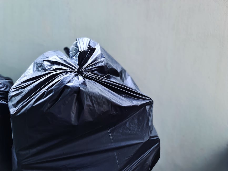 A person's hand and forearm extend into the frame from the left side, grasping a green plastic rubbish bag by its twisted top. The bag appears filled with lightweight waste material, exhibiting bulges
